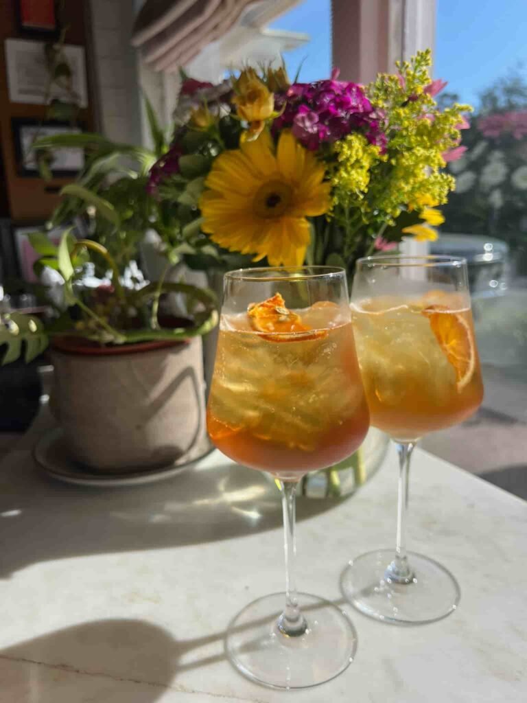 Two elegant wine glasses filled with a golden-hued cocktail, garnished with dried orange slices, sit on a sunlit marble countertop. A vibrant bouquet of yellow, pink, and green flowers in the background adds a fresh, cheerful touch. The scene is bathed in warm natural light, enhancing the inviting ambiance.