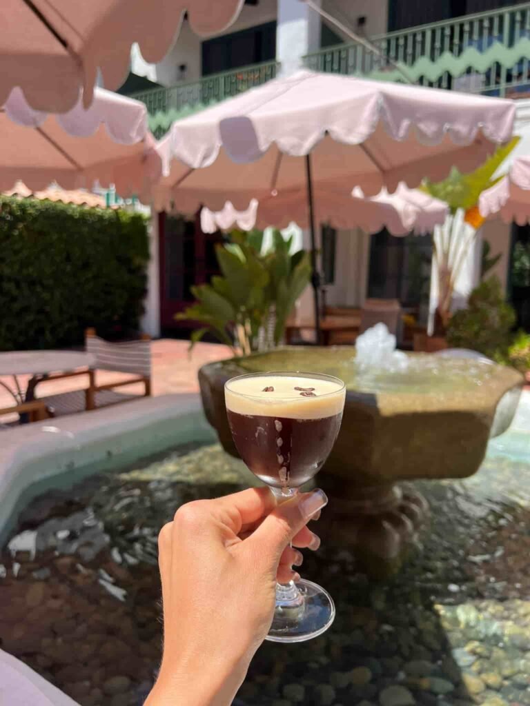A hand with neatly manicured nails holds a small glass of espresso martini garnished with three coffee beans. The background features a courtyard with a stone fountain, a shallow pool of water, and pink scalloped patio umbrellas providing shade. Green plants and a Mediterranean-style building with wrought iron balconies add to the relaxed, elegant atmosphere.