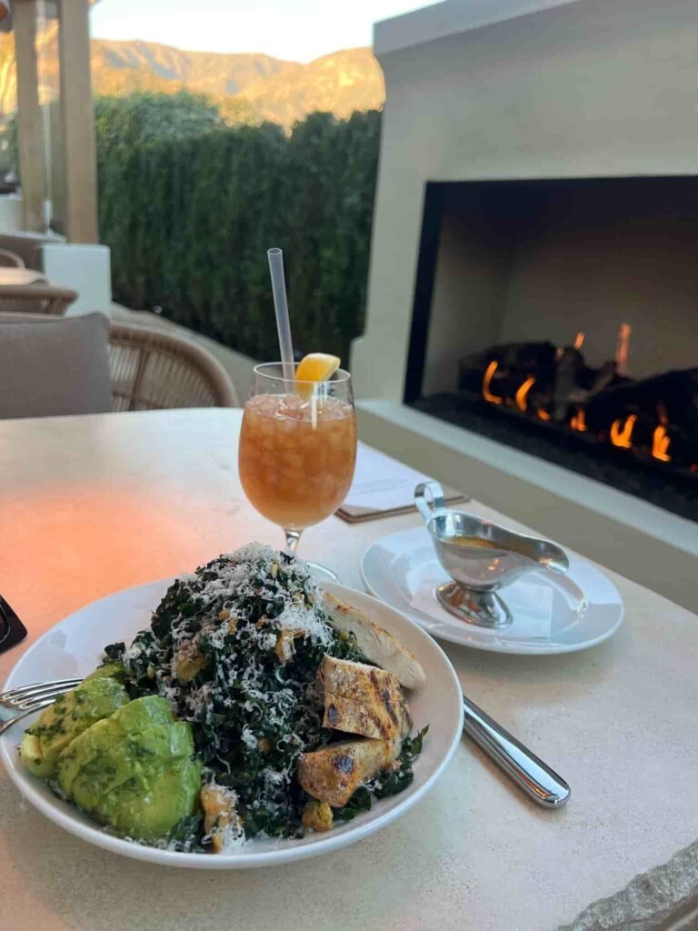A fresh kale salad topped with grilled chicken, avocado slices, croutons, and shredded cheese is served on a white plate. A tropical cocktail with crushed ice and a lemon garnish sits beside it, with a small silver sauce boat on a separate plate. In the background, a cozy fireplace glows, and mountains are visible beyond a lush green hedge.